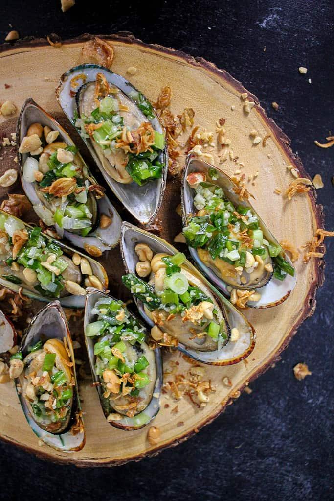 Five Grilled Scallion Oil Mussels (Chem Chép Nướng Mỡ Hành) arranged on a board that looks like a slice of a tree trunk. Fried shallots are strewn on top of the board.